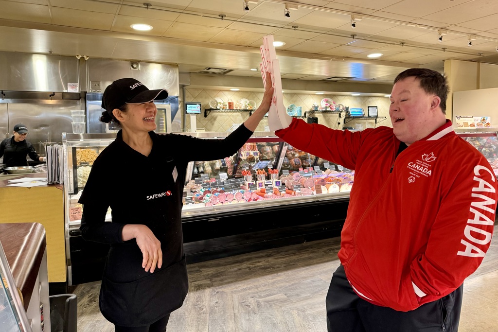 A Special Olympics Saskatchewan athlete is high-fiving a Safeway employee. They are both smiling and happy. The Safeway employee is wearing her black uniform while the athlete is wearing a red Canada sweater and a white foam hand.