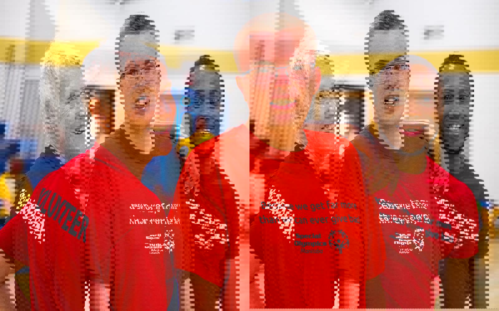 Three people smiling for the camera. The woman on the left has her back to the camera to show that her red shirt reads "VOLUNTEER", and she is looking over her shoulder. The athlete on the right has his arm resting on the shoulder of the athlete in the middle.