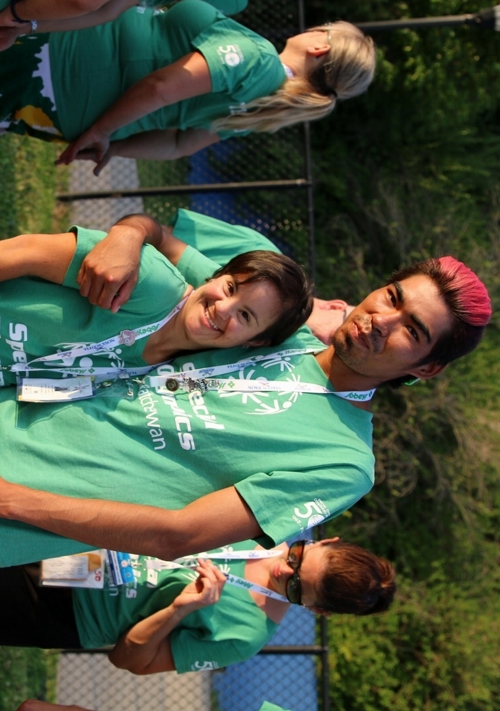 Two Special Olympics Saskatchewan athletes are wearing matching green "Special Olympics Saskatchewan" t-shirts and smiling for the camera. They have their arms wrapped behind each other and they are standing on a track.