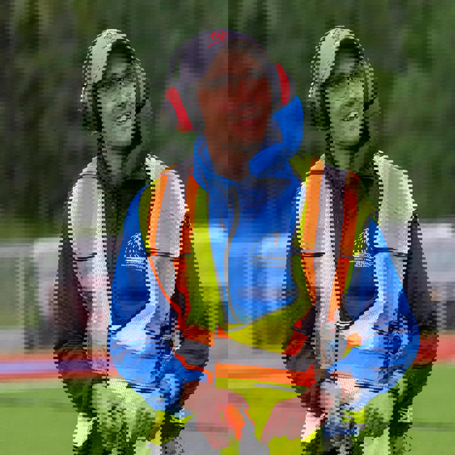 An image of a race starter, on his knees, reloading the starting gun. He is wearing a high visibility vest as well as large red headphones to protect his ears. He is looking up and to the right of the camera, smiling and listening to someone who does not appear in the photo.