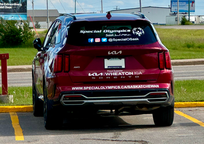 A red Kia car is parked in a parking spot. The car has the Special Olympics Saskatchewan logo on the window along with the social media icons and social media handle in white letters. Also in white letters and written along the bottom of the car is Special Olympics Saskatchewan's website.