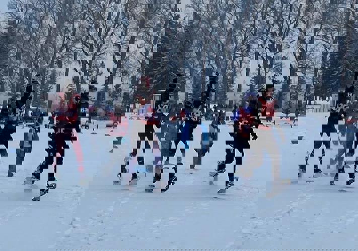 An action shot image of snowshoers. There are five athletes running across the snow a medium distance away from the camera. In the distance behind them, you can see trees and viewers watching the sport.