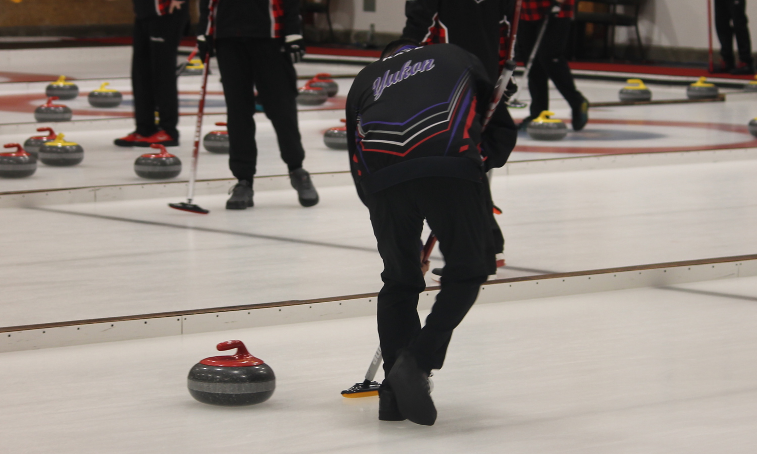 An action shot photo of an athlete curling. You can only see them from behind. They are wearing a black jacket that has "Yukon" written on the back of it. They are using the curling stick to direct the curling stone across the ice.