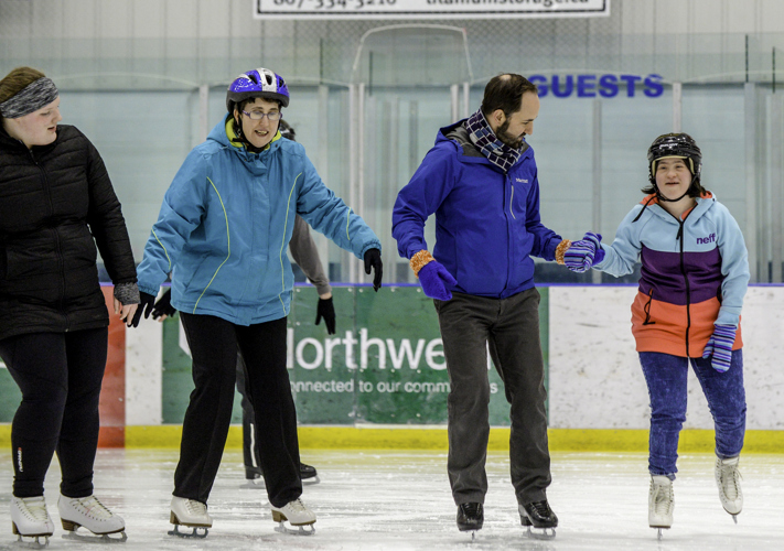 Five figure skaters of all ages skating on the ice. They are all wearing winter jackets of different colours (from left to right: black, light blue, dark blue, blue/purple/pink, and pink). Some skaters are also wearing helmets of different colours (from left to right: black headband, blue helmet, no helmet, black helmet, pink helmet). Each athlete is focused on their skating.