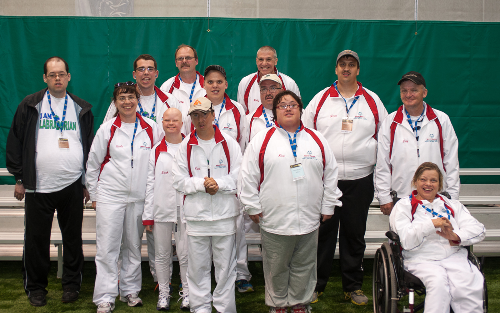 A team photo. Everyone is wearing white tracksuits and blue lanyards around their necks. They are standing in an indoor field.