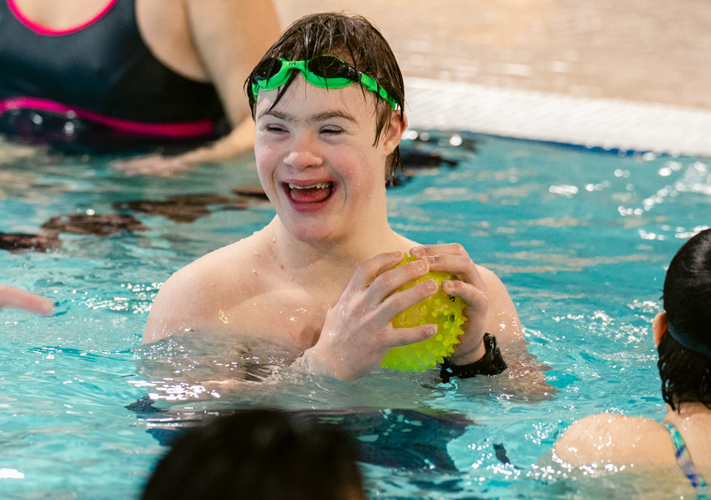A swimmer happily swimming in a pool holding a yellow spiky ball. He has green googles on his forehead and looking at someone not pictured, but their arm is seen pointing at something. There are two more swimmers in the photo but you can only see their backs.