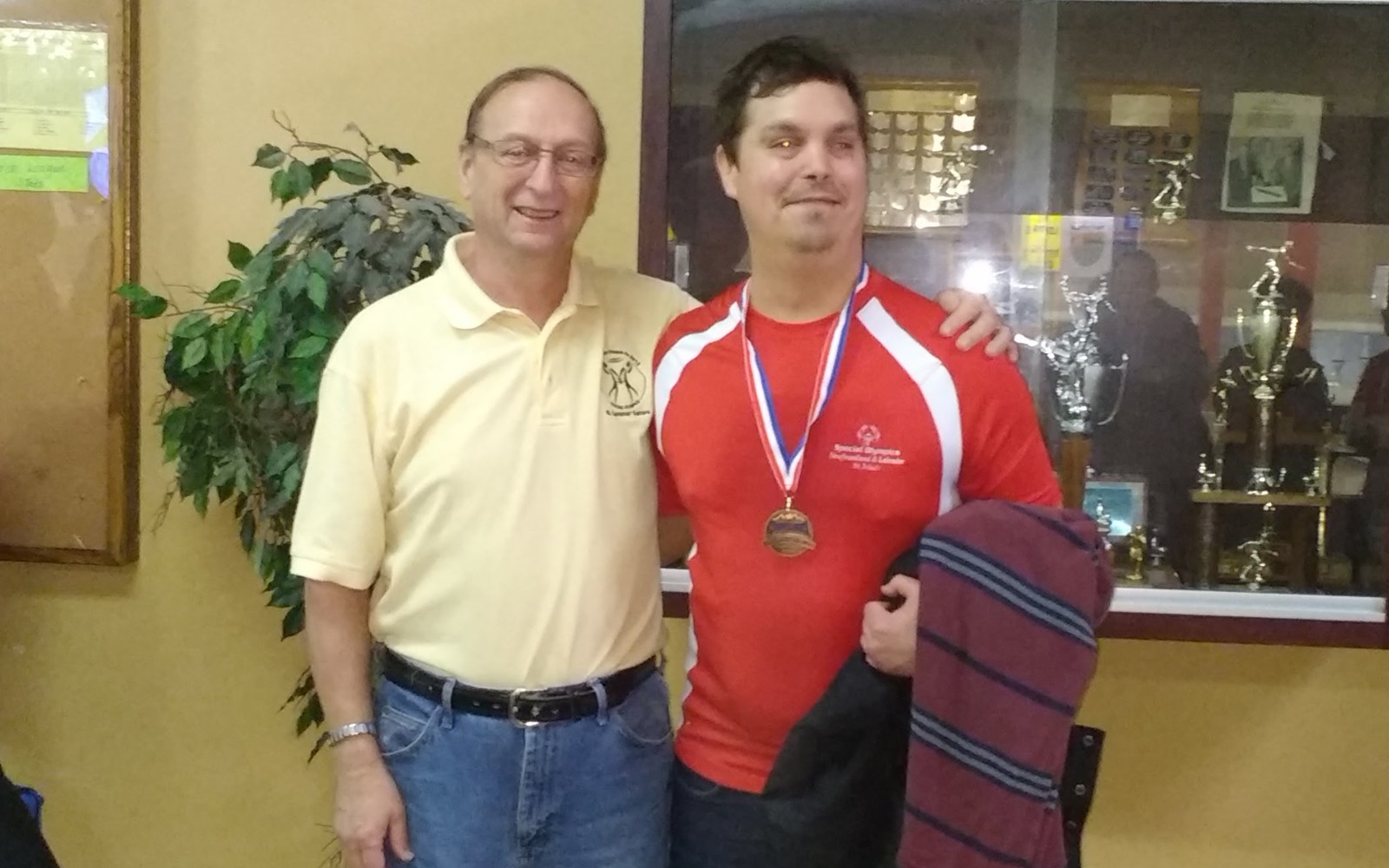 Two individuals are posing and smiling for the camera. The athlete on the right is wearing a red jersey and has a medal around his neck. The individual on the left is wearing a yellow collared t-shirt and has his arm around the back of the athlete. They are standing in a room, and are in front of a trophy case.