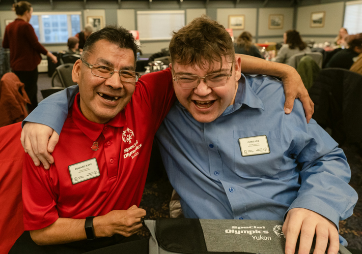 Two athlete ambassadors have their arms wrapped behind each other and smiling for the camera. They are sitting at a table during a formal event. The athlete ambassador on the left is wearing a red t-shirt while the athlete ambassador on the right is wearing a blue long sleeved button up dress shirt.