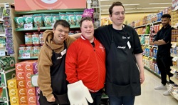 A Special Olympics athlete poses and smiles for the camera with two Sobeys workers. They are standing in a Sobeys grocery store, in front of one of the aisles. The Sobeys employees are wearing their black "Sobeys" aprons while the athlete is wearing a red sweater and a white foam hand.