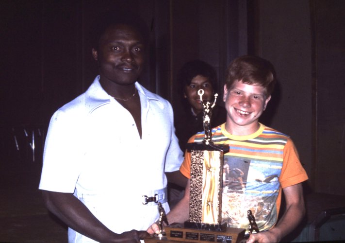 A young athlete in an orange t-shirt is shaking hands with a man wearing a white t-shirt awarding him with a trophy. They are both smiling and posing for the camera.