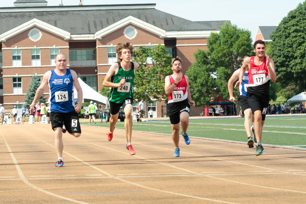 Four athletes are running on the track in the middle of a race. The athletes are all wearing different coloured jerseys. From left to right the coloured jerseys are; blue, green, and two in red. All the athletes are focused on their match.