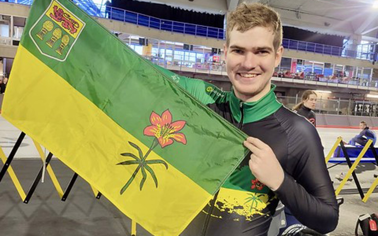 A picture of Jordan, a Special Olympics Saskatchewan athlete holding up a Saskatchewan flag. He is standing in an ice rink but next to the ice.