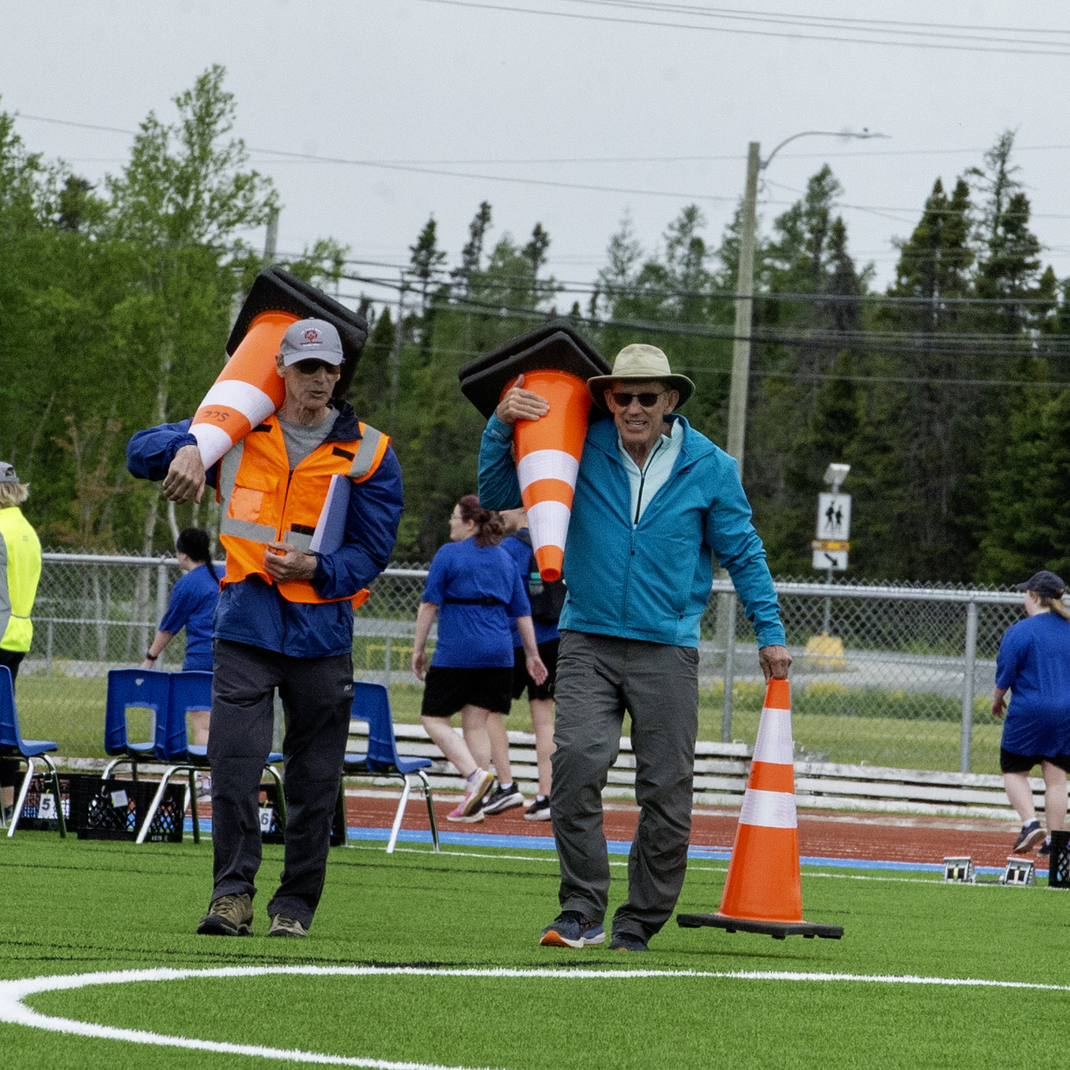 Two individuals are walking on a field holding large, orange, pylons. The individual on the left is wearing a high visibility vest and is holding one pylon while the individual on the right is wearing a blue jacket and holding two pylons. In the background you can see other officials and volunteers in a group.