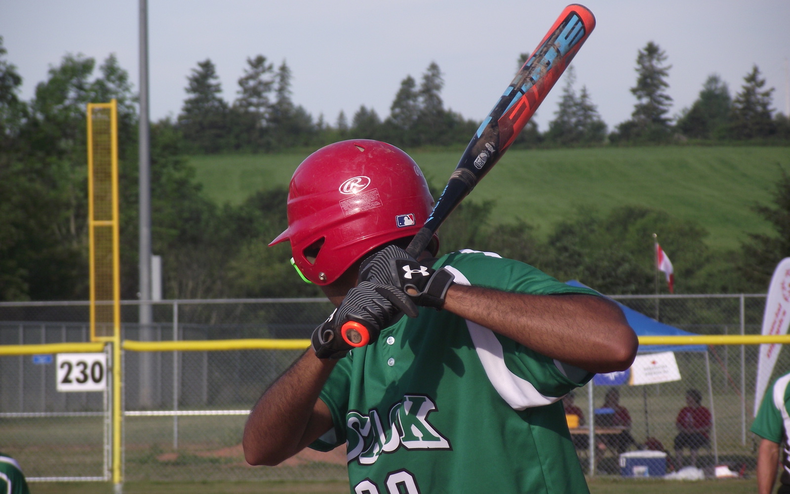 A close up image of an athlete playing baseball. They have their face turned away from the camera as they are focusing on their game. They are swinging the baseball bat back in preparation to hit the ball. They are wearing a green baseball jersey.