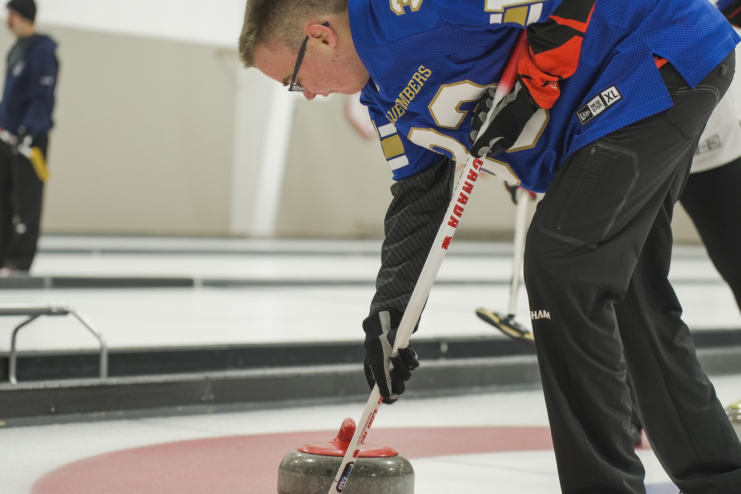 A close up image of a curling athlete. The athlete is holding his curling stick in front of the curling stone. He is wearing a blue jersey.