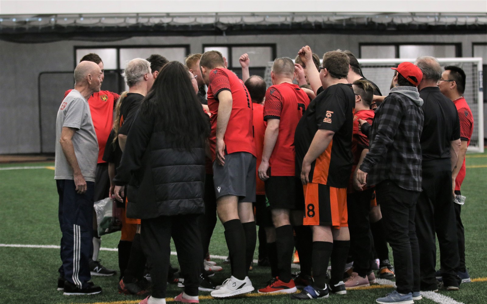A large soccer team is in a huddle together. This includes athletes in their soccer uniforms as well as coaches and volunteers. We can only see everyone's backs. Some of the athletes have their hands raised in fists, participating in the group's cheer.