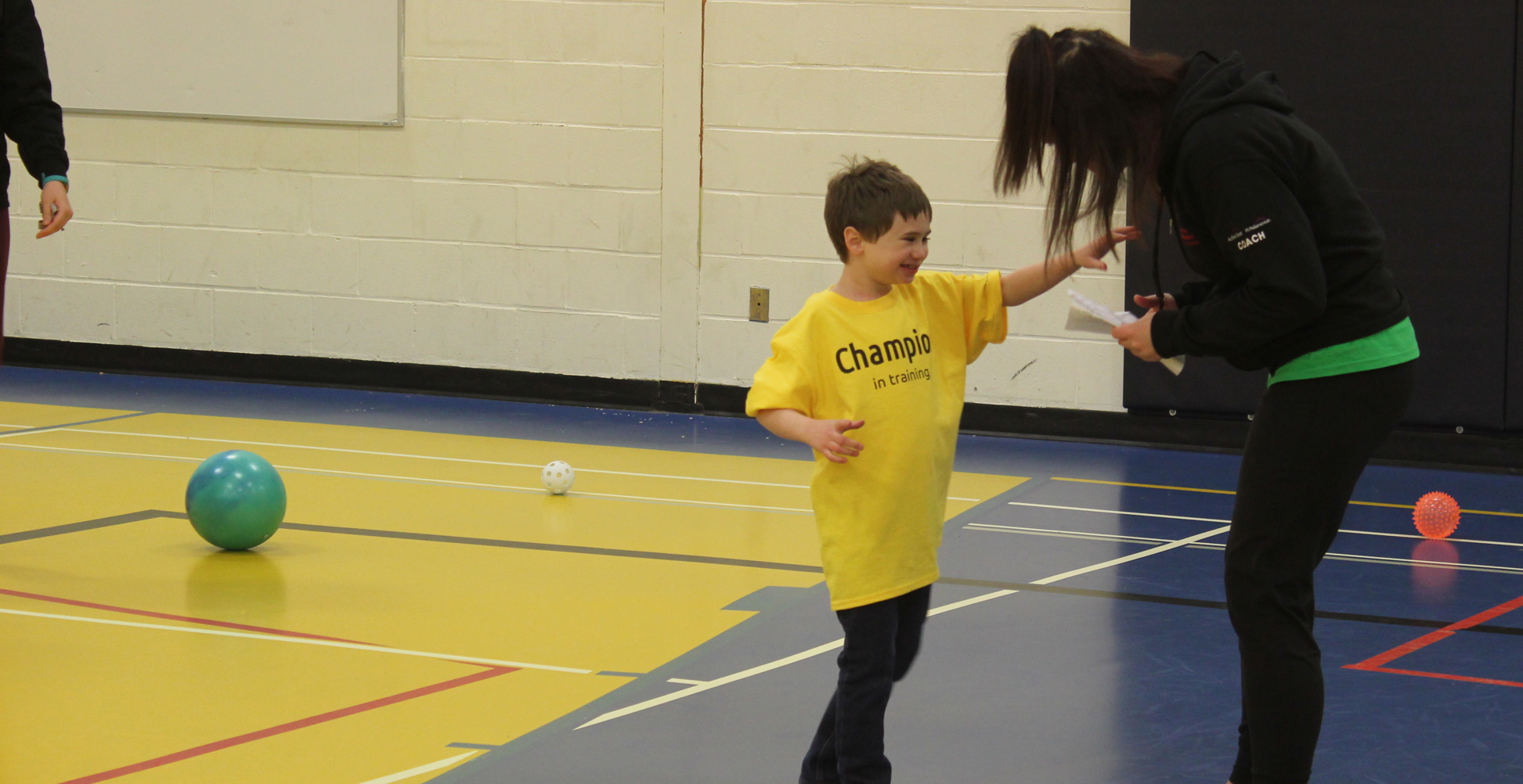 A young athlete in a yellow t-shirt that says "Champion in Training" on it, is reaching his arm out towards his instructor. They are both in a gymnasium and different coloured balls are scattered across the floor.