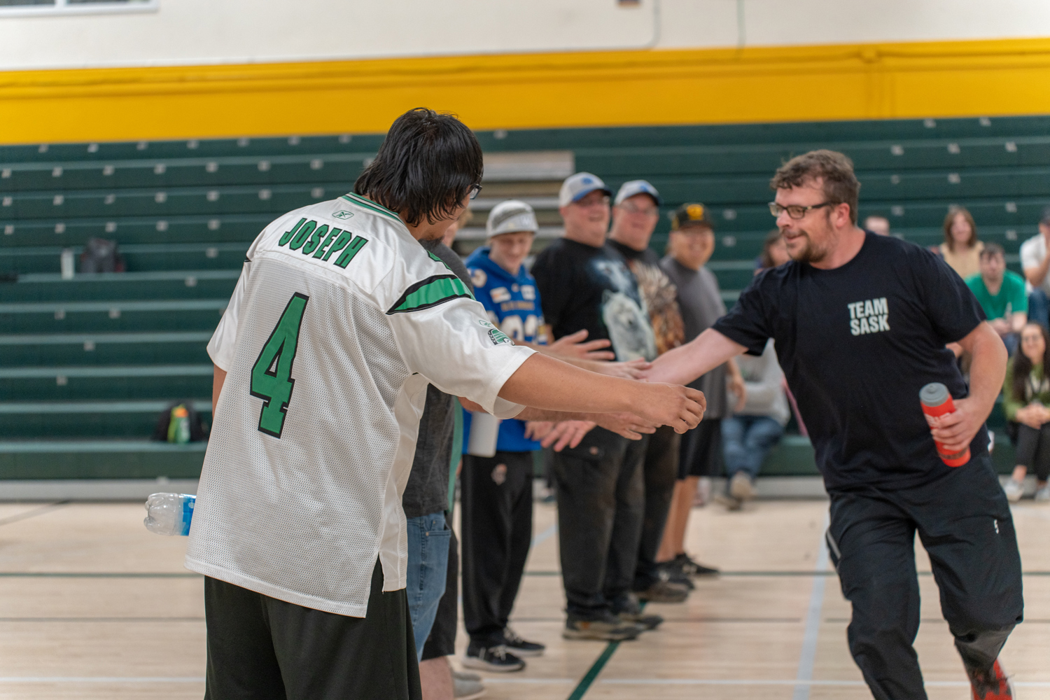 A man wearing a black t-shirt that says "Team Sask" on it, is walking down a lineup of athletes and giving them a high-five. The athlete in the front of the line is in the focus of the camera. He is wearing a white and green jersey with "Joseph" and the number "4" on the back. Everyone is in a gymnasium.