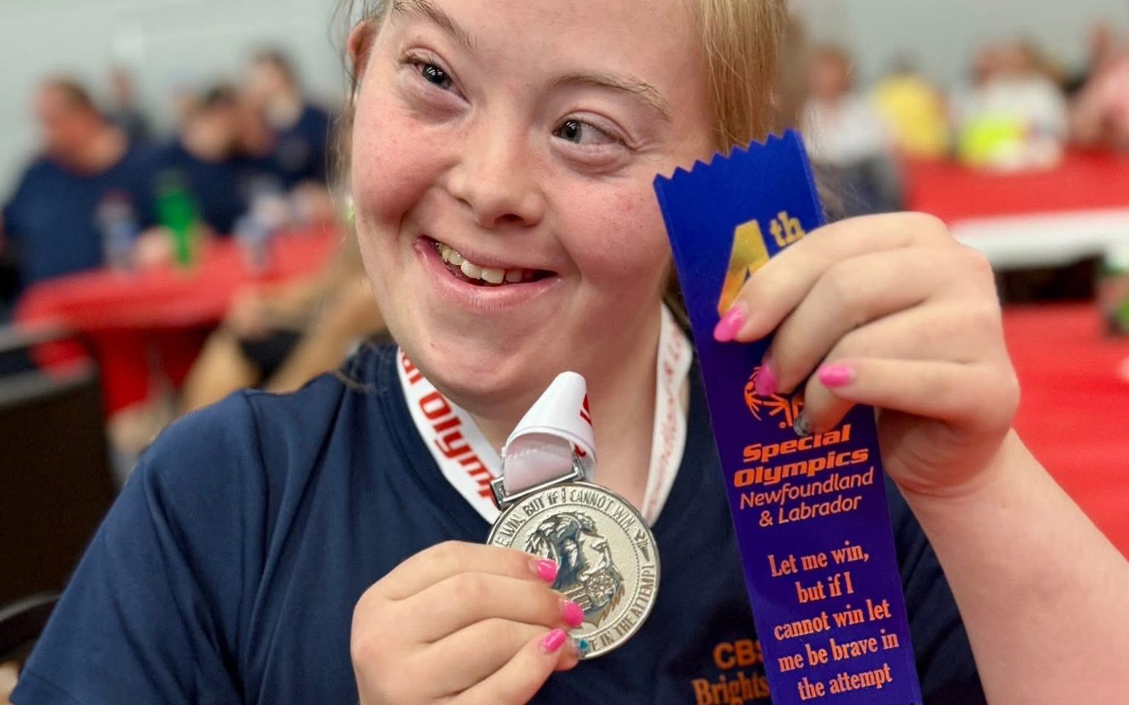 A close up image of an athlete holding up the medal and ribbon she won. The silver medal is hanging around her neck, and she is holding up the medal in one hand. In the other hand is a blue 4th place ribbon. The athlete is wearing a navy blue t-shirt with the Special Olympics Newfoundland & Labrador CBS Brightstars logo on it.