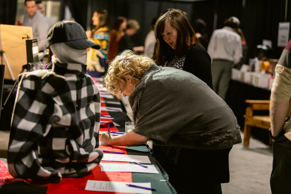 The photo is showing an auction. On the table is half a mannequin with a plaid shirt and a baseball cap on. Two women are looking at the table that has forms and pens on it. One woman is writing on one of the forms. She is sideways to the camera.