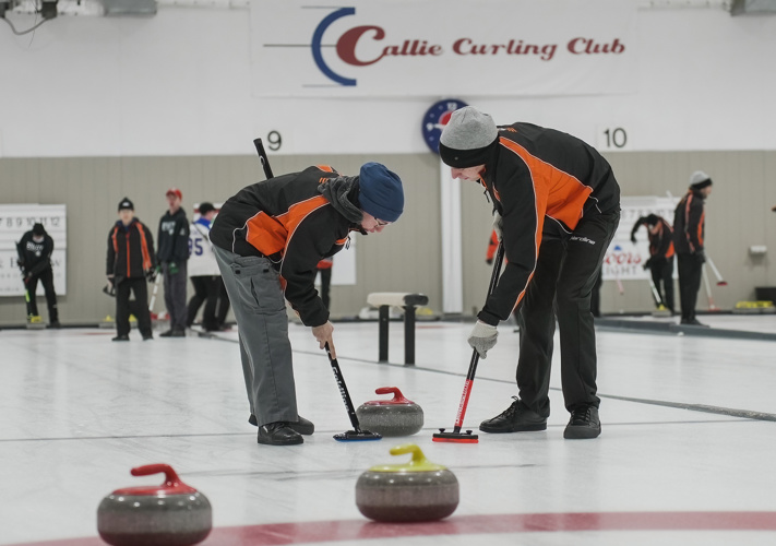 An action shot of a curling game. Two curling athletes are using their curling sticks to help the curling stone slide across the ice. They are wearing black and orange jackets and are focused on the game.