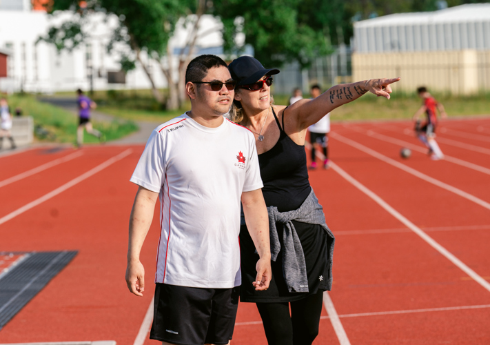 A coach is standing behind an athlete and pointing to the side, giving direction to the athlete. They are standing on a track. The coach is wearing all black with a black baseball cap. The athlete is wearing a white t-shirt and sunglasses.