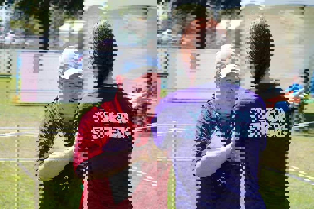 A volunteer wearing a purple shirt that says "Yukon Strong", assisting an athlete. The athlete is wearing a red t-shirt and grey baseball cap, and pointing next to the volunteer.