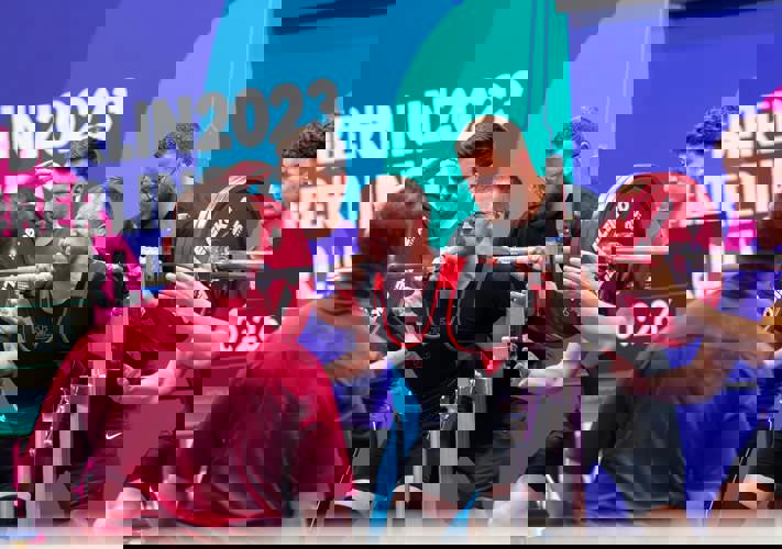 Young man gritting his teeth to lift a heavy barbell. Other people stand nearby to help as needed. 