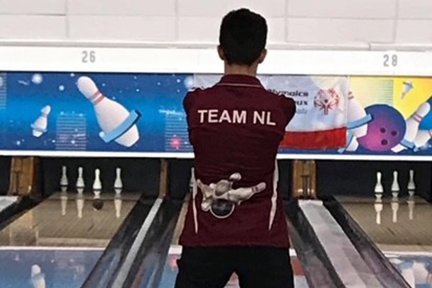 A bowling athlete is standing at the top of the bowling aisle, preparing for their turn. The camera is directly behind the athlete. The athlete is wearing a burgundy "Team NL" bowling shirt.