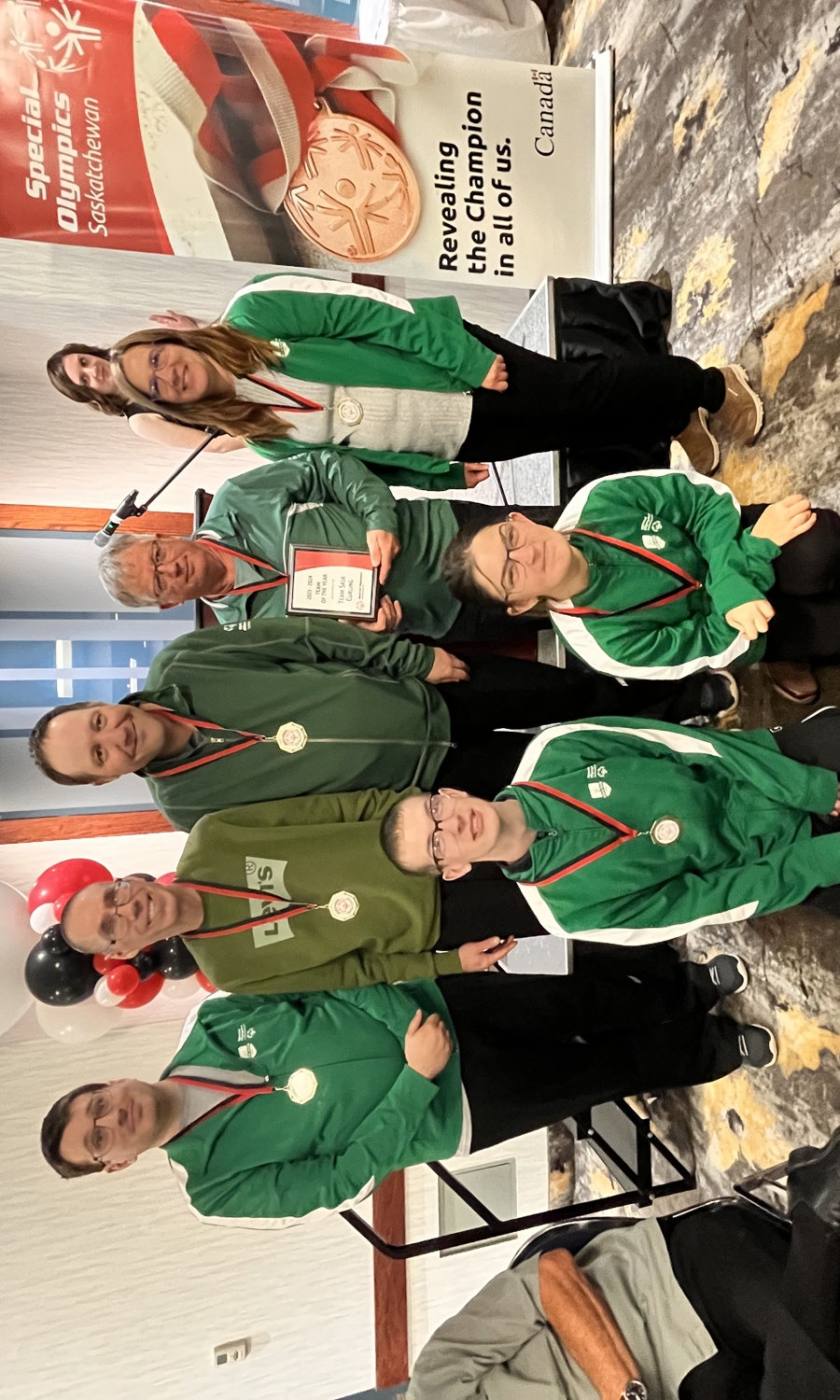 A group team photo of athletes who have won medals. They pose, two people in front on their knees and five standing behind them. They are all wearing green sweaters with a ribboned medal hanging around their necks. The image takes place in a large room with banners and balloons behind the team.