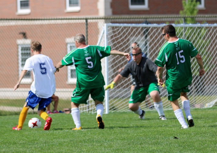 An action shot photo of a soccer match. Four athletes can be seen on the soccer field playing soccer. One player in a white jersey has possession of the soccer ball and is approaching the net. The goalie in the net has his arms out ready to guard the net. Two players in green jerseys are defending the net and running towards the player in possession of the ball.