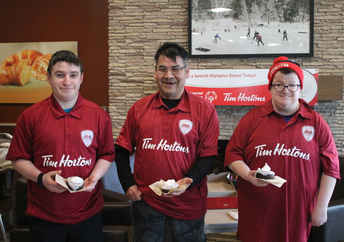 Three athletes smile and pose for the camera holding a Special Olympics donut from Tim Hortons. They are each wearing red Tim Hortons shirts and they are standing inside a Tim Hortons shop.
