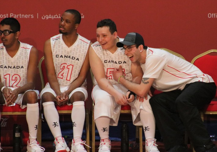 4 men sit on a bench. They are all dressed in white and red Team Canada basketball uniforms. One of the men is leaning over and cheering. 