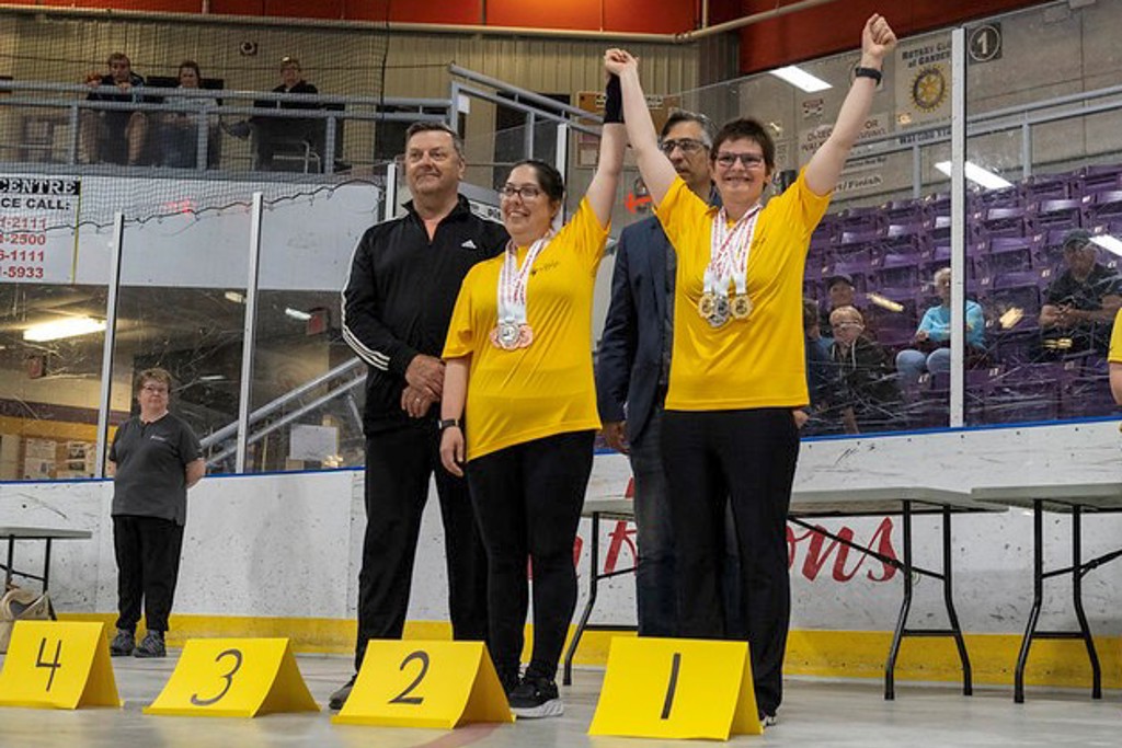 Two winning athletes are standing next to each other holding hands, with their arms raised in the air. They are both wearing yellow t-shirts and have multiple medals hanging around their necks. There are paper signs in front of them that read "1", "2", "3", "4" from right to left.