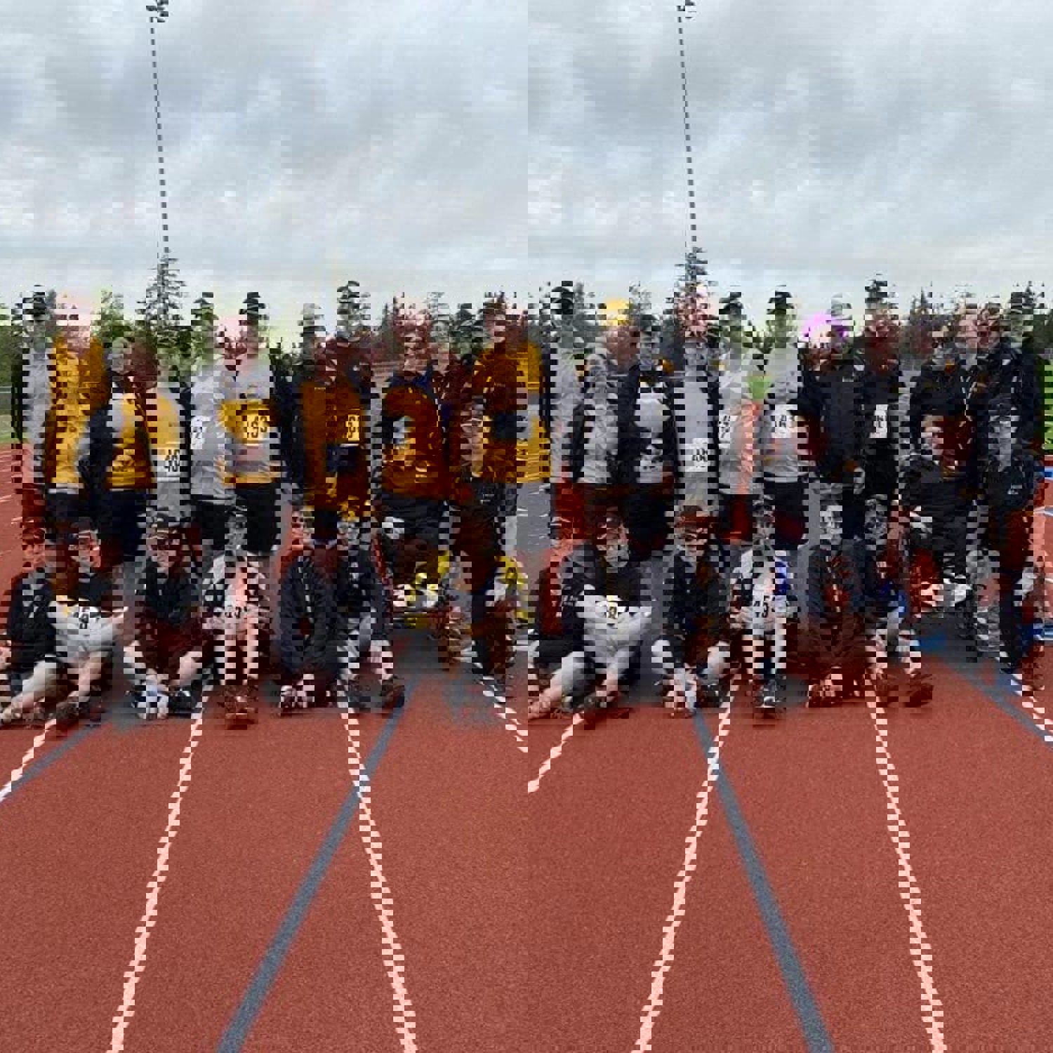A group, team photo on the track. Everyone is in matching yellow and black team uniforms. The front row of people are sitting on the track while the back row of people are standing.