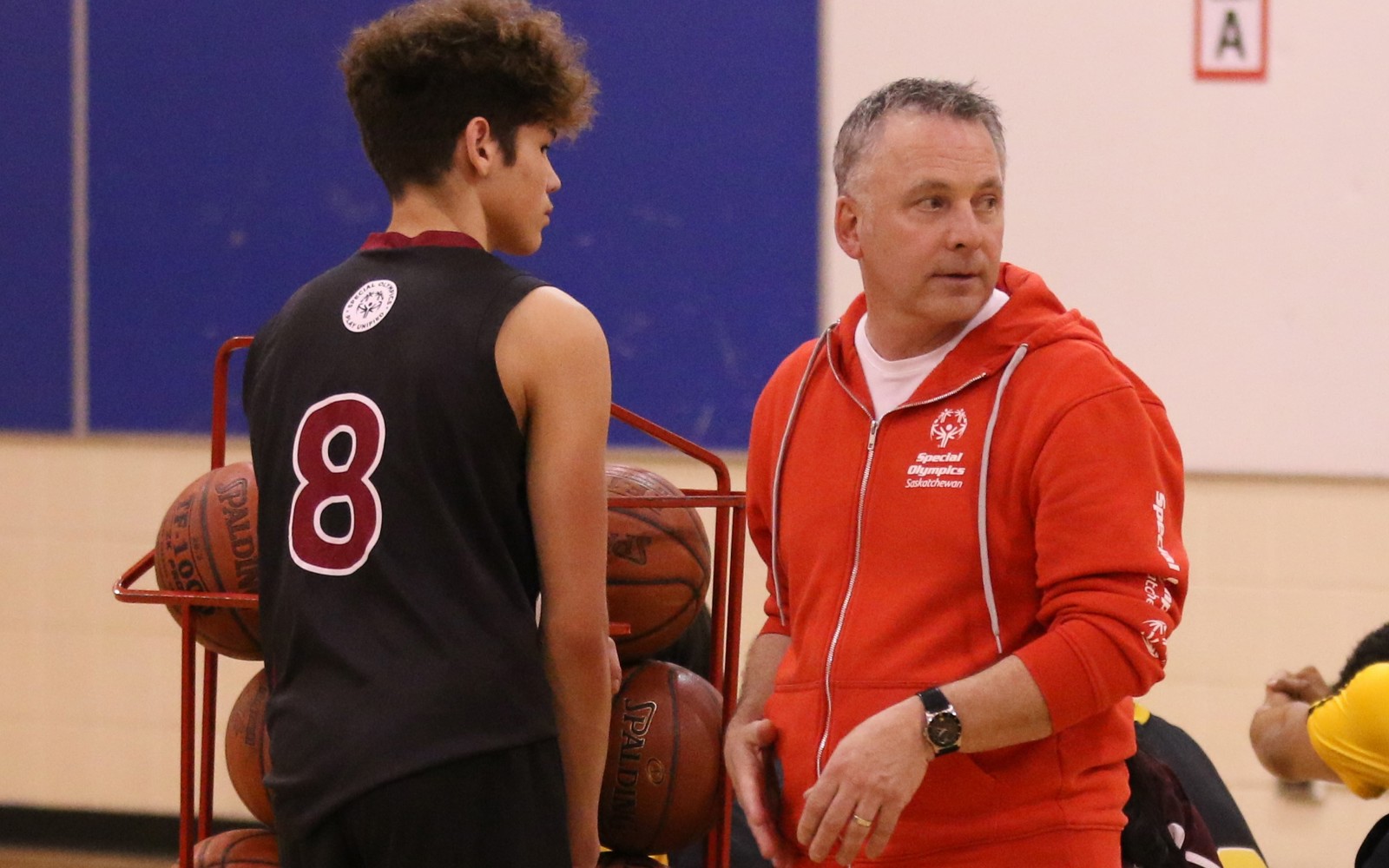 A basketball coach talking to a basketball athlete. They are standing to the side of the court next to a rack filled with basketballs.