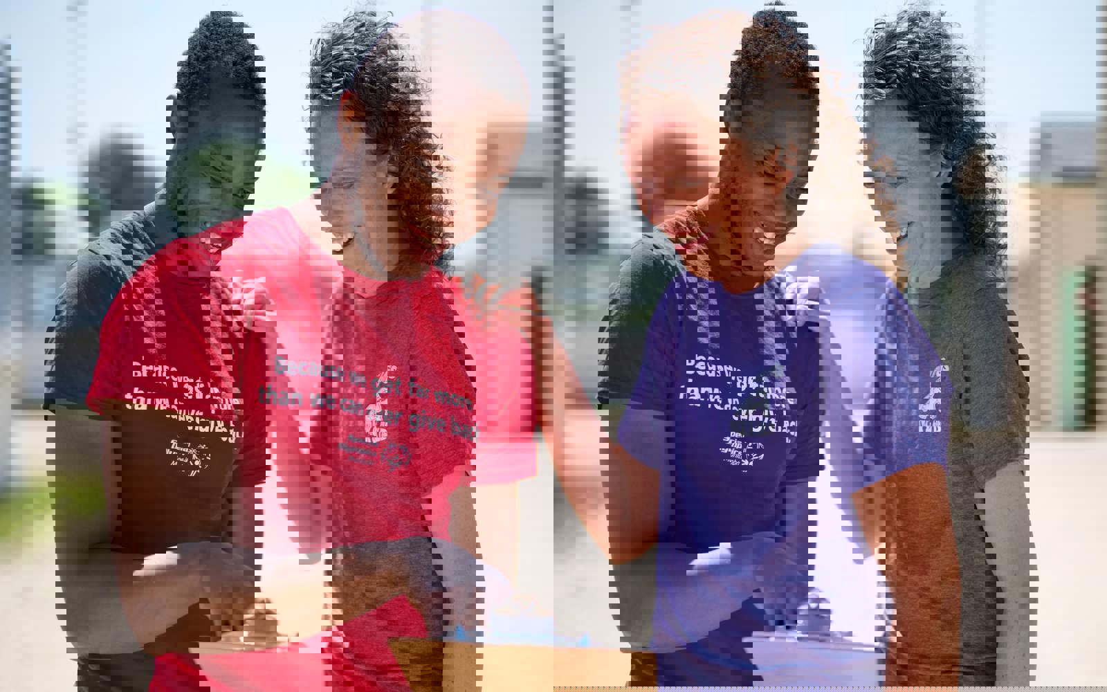 A person in a red shirt with the words "Because we get far more than we can ever give back" holding and pointing at a clipboard while a person in a purple shirt smiles down at the clipboard. 