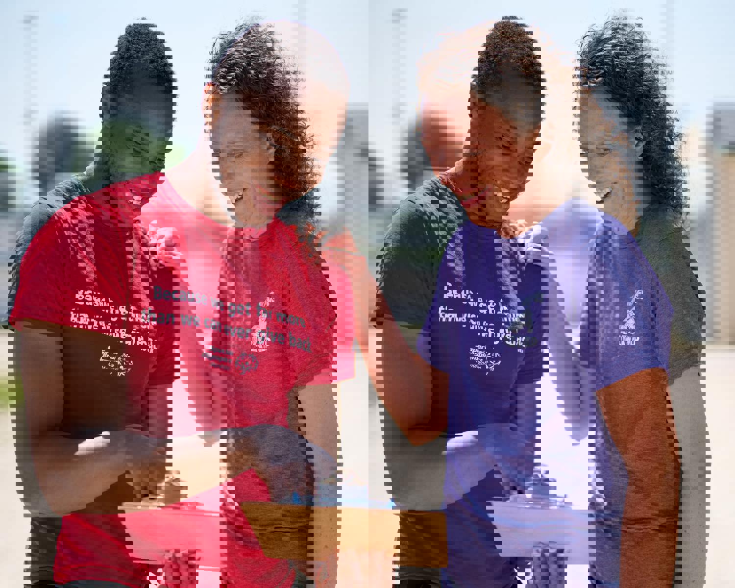 A person in a red shirt with the words "Because we get far more than we can ever give back" holding and pointing at a clipboard while a person in a purple shirt smiles down at the clipboard. 