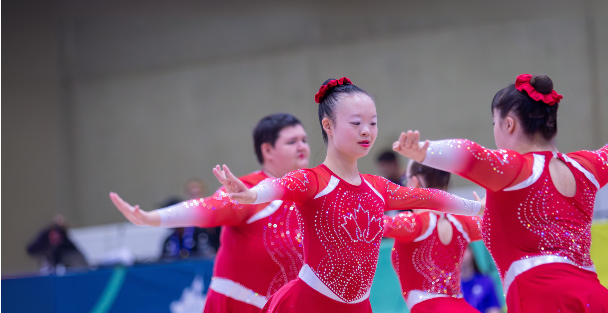Diverse group of people in red outfits with the maple leaf in sparkles. They appear to be practicing figuring skating. 