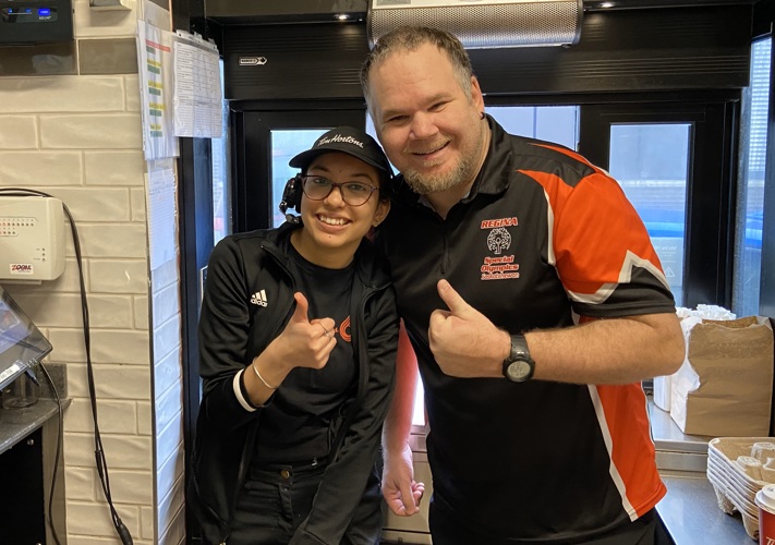 A Special Olympics Saskatchewan athlete is posing with a Tim Hortons employee at the drive through window. They are both giving a thumbs up and smiling for the camera.