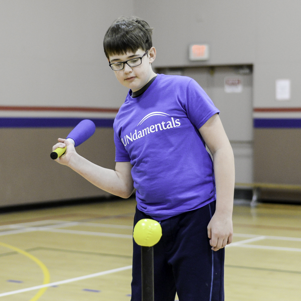 A young athlete in a blue FUNdamentals t-shirt, is swinging back a baseball bat to hit a yellow ball perched on a post. He is in a gymnasium and wearing a pair of glasses.