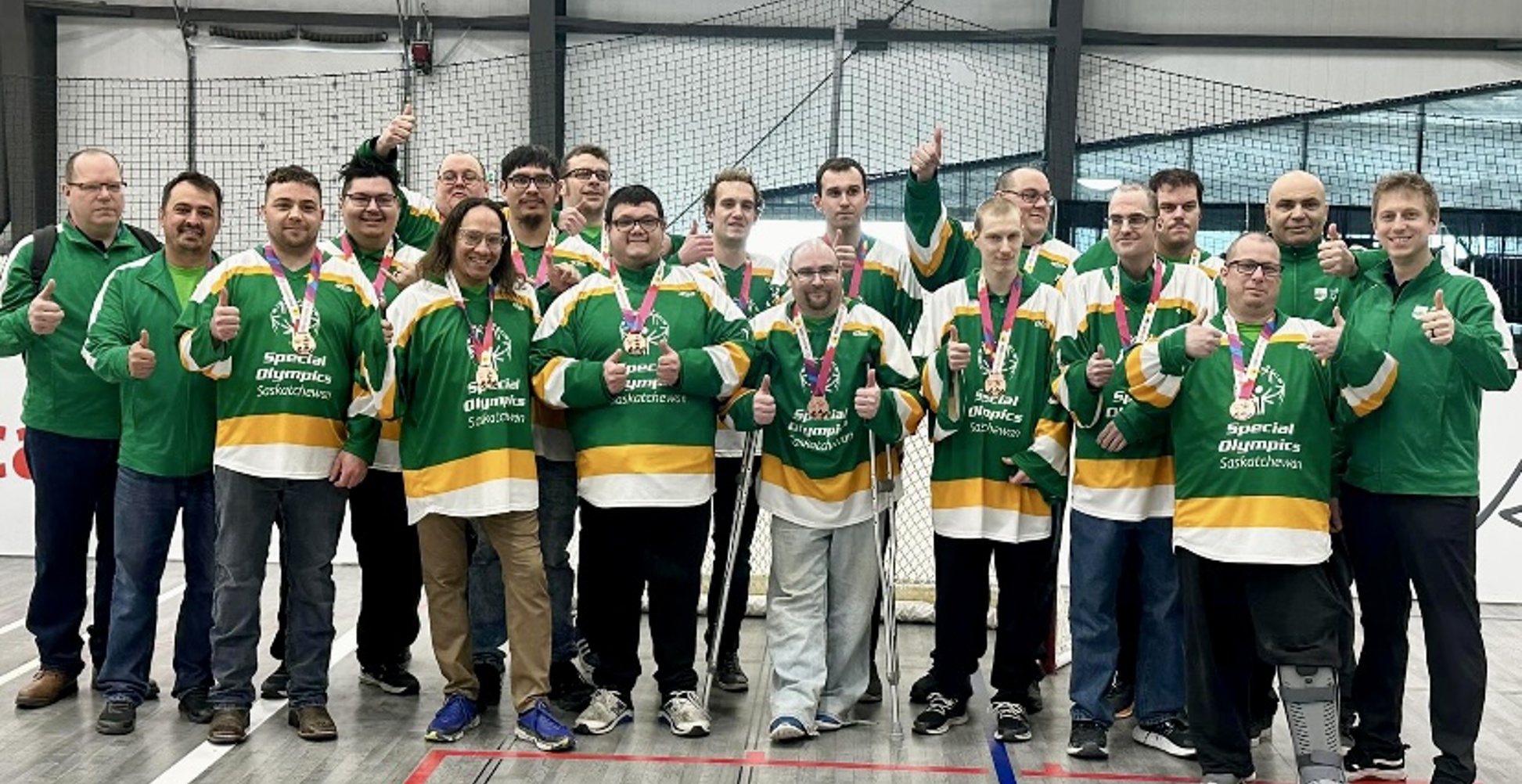 A group photo of the Special Olympics Saskatchewan Floor Hockey Team. There are 19 men in the photo, all smiling and giving thumbs up to the camera. The athletes are wearing green jerseys with medals around their necks.