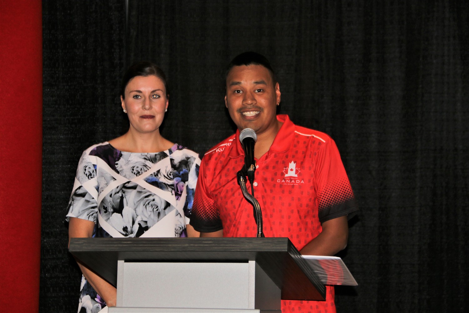 Two individuals are standing behind a podium and a microphone and giving a speech. They both look very happy to be giving their speech.
