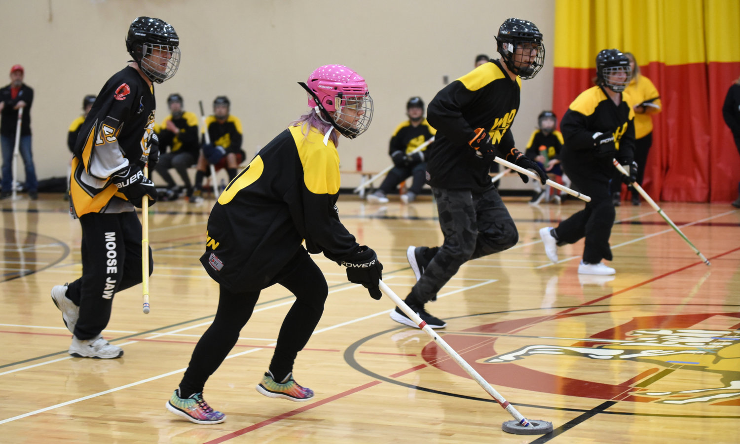 An action shot photo of athletes playing floor hockey. Four athletes can be seen running across the gym floor with their floor hockey sticks. They are all wearing helmets and black and yellow floor hockey jerseys.