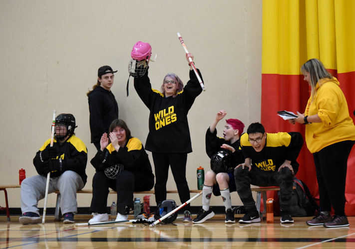 Floor hockey teammates are sitting on the bench and cheering for their teammates. Everyone is smiling, has their hands up, and are clapping for their teammates.