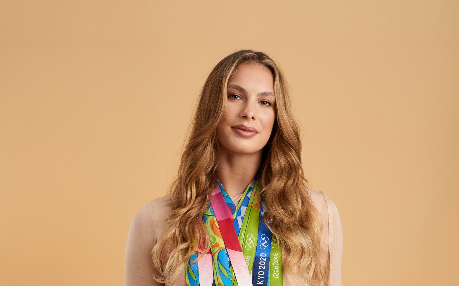 Headshot of a women in a beige dress with long blond wavy hair. Around her neck is many Olympic medals. 
