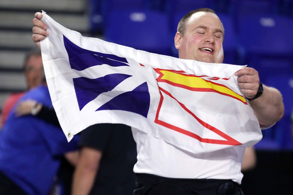 An athlete wearing a white t-shirt and thick red belt, is holding up the Newfoundland flag. He is smiling with his eyes closed. He is in clear focus as the background around him is blurred.