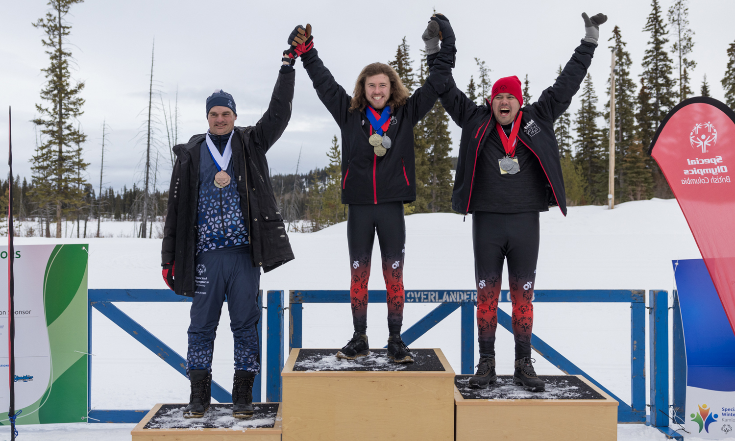 Three athletes are standing on a three tiered podium. They are all holding hands and lifting them in the air. Around their necks are the medals they have won and each athlete is smiling.
