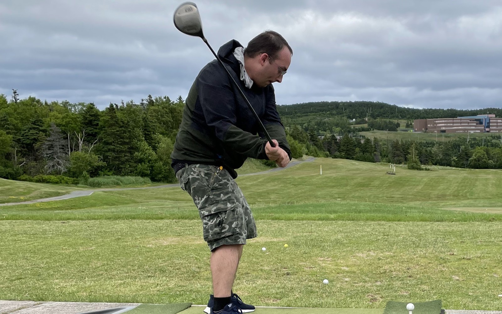 An action shot photo of a golf athlete taking a swing at the start of the golf game. It is a cloudy day and the athlete is wearing a black sweater. In the distance you can see the other golf balls that have been in play.