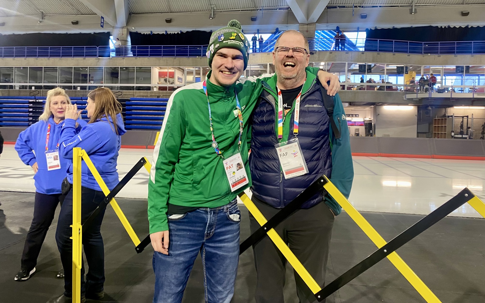 An athlete posing with his coach after the game. They are both smiling for the camera with their arms wrapped behind each other. The athlete is wearing a green sweater, a pair of jeans, and a winter hat. The coach is wearing a navy vest, and a teal sweater. They both have a lanyard around their necks.